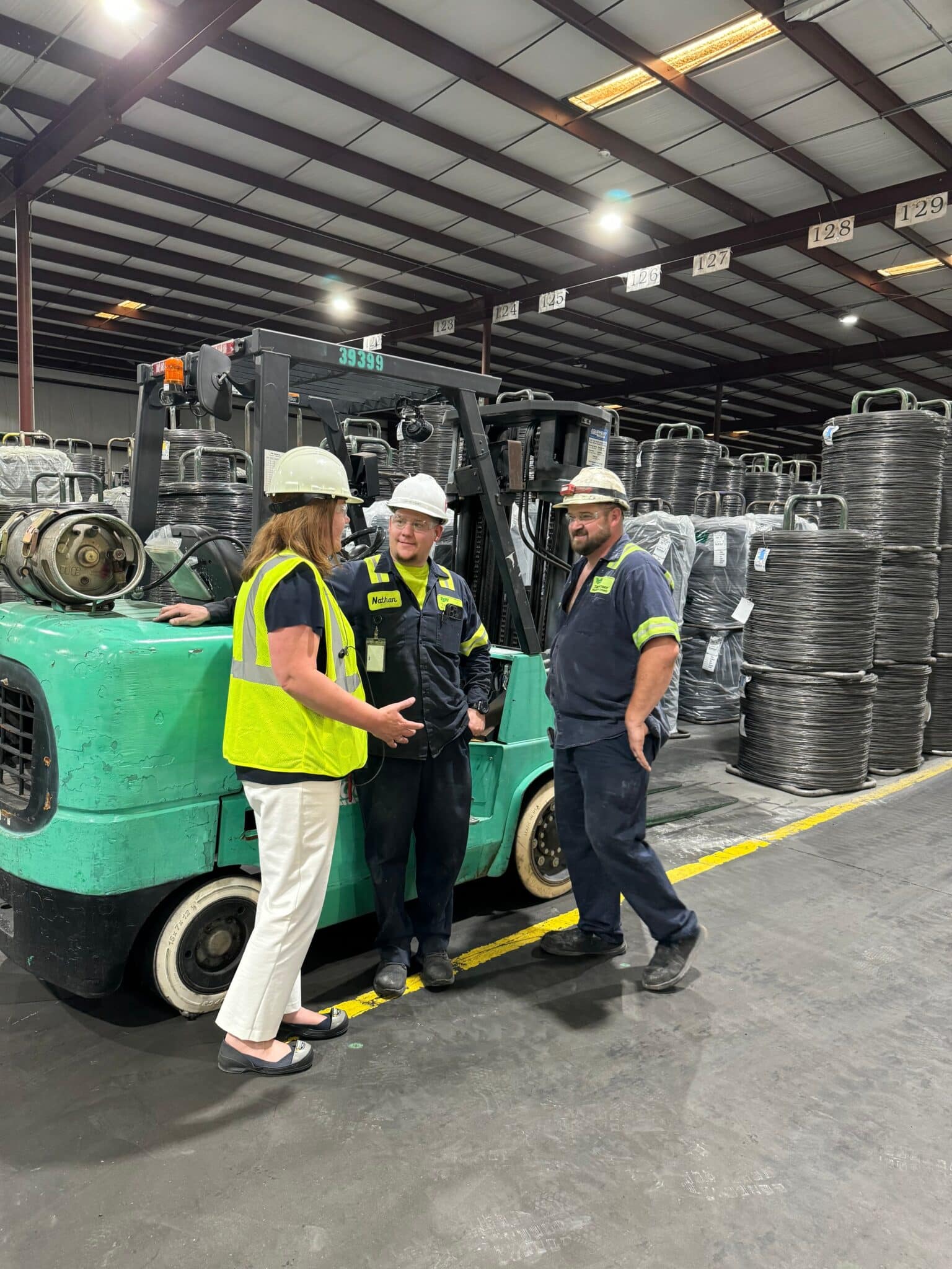 Three people from the Taubensee team discussing in a warehouse, all wearing hard hats. They are standing next to a forklift and stacks of low carbon wire.